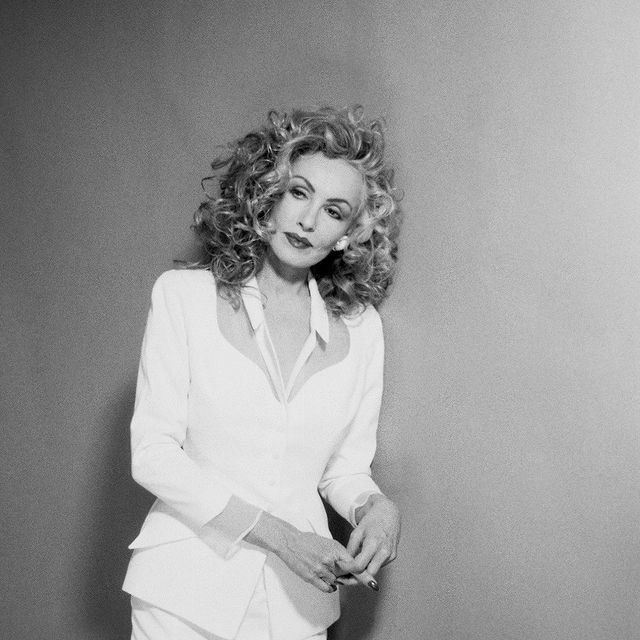 Julie Newmar standing against a wall wearing a white shirt in her curly hair.